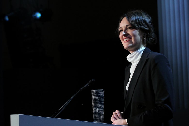 Phoebe Waller-Bridge stands at a podium in a dimly lit room.